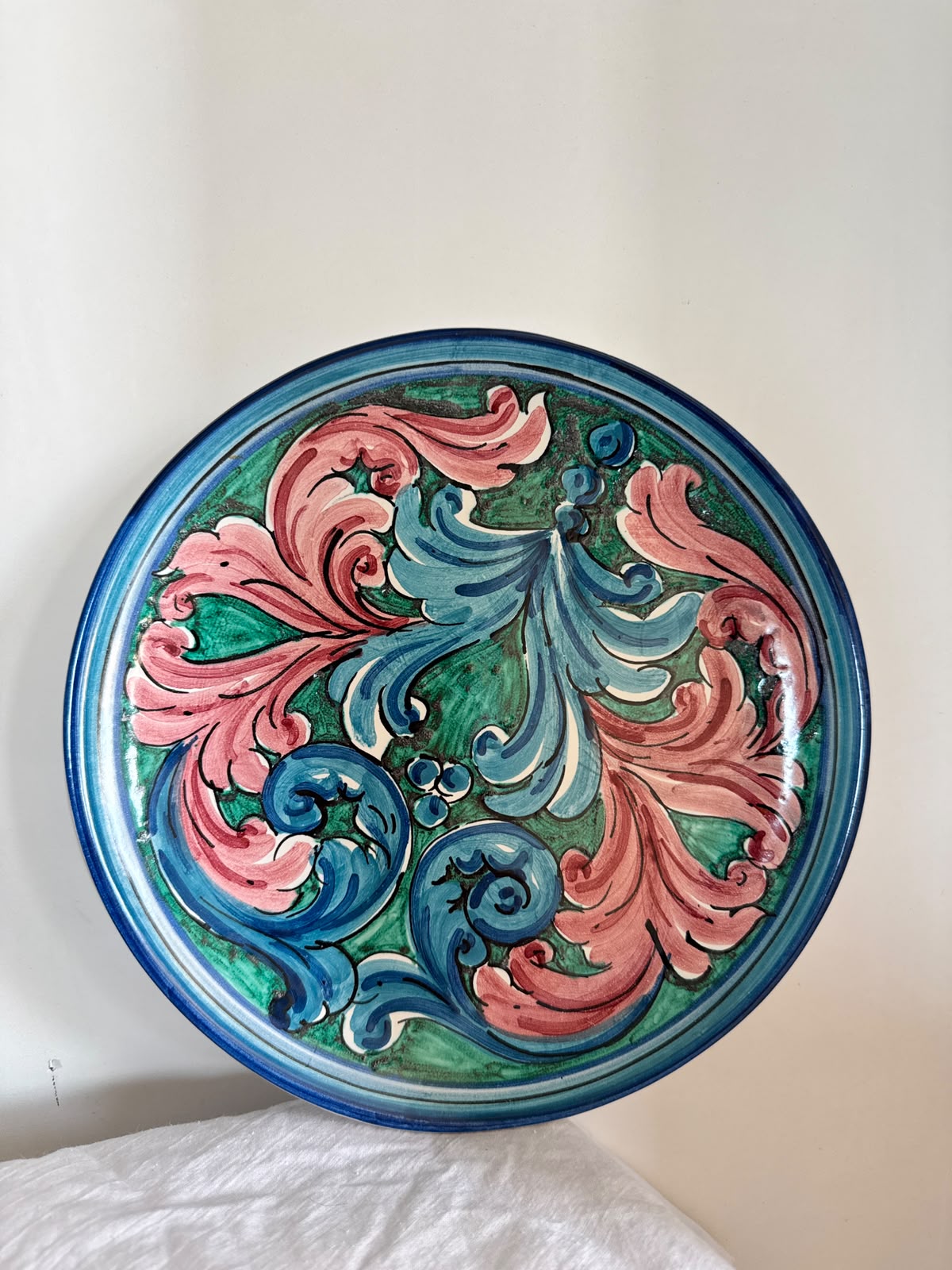 Hand-painted Sicilian ceramic decorative plate with pink, blue and green baroque motifs, displayed on natural white linen to highlight its artisan details.