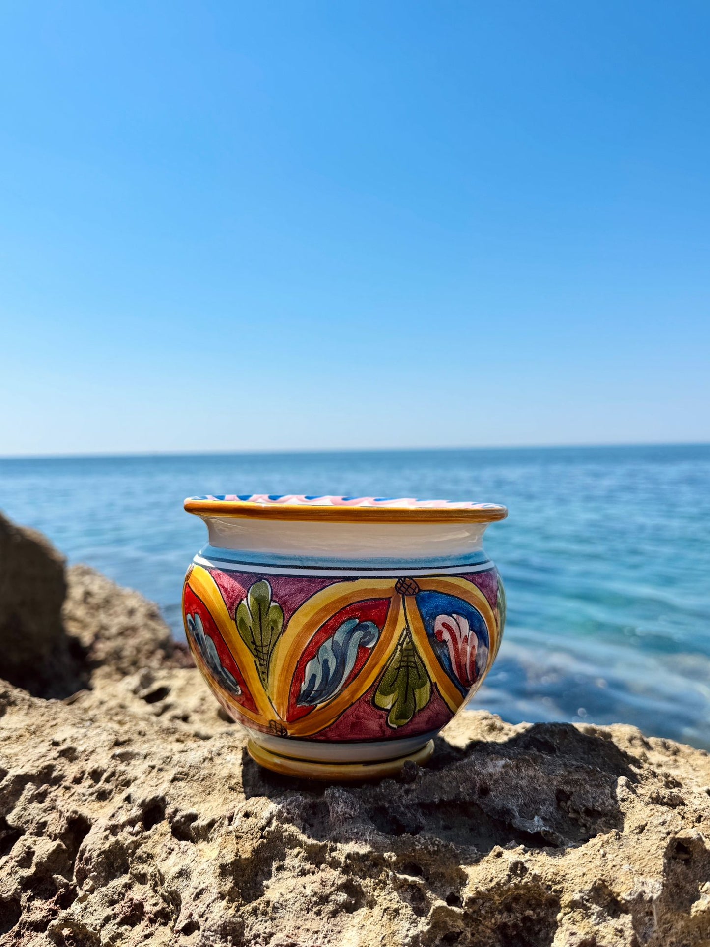 Close-up of a handcrafted Sicilian ceramic cachepot resting on seaside rocks with the Mediterranean Sea in the background.