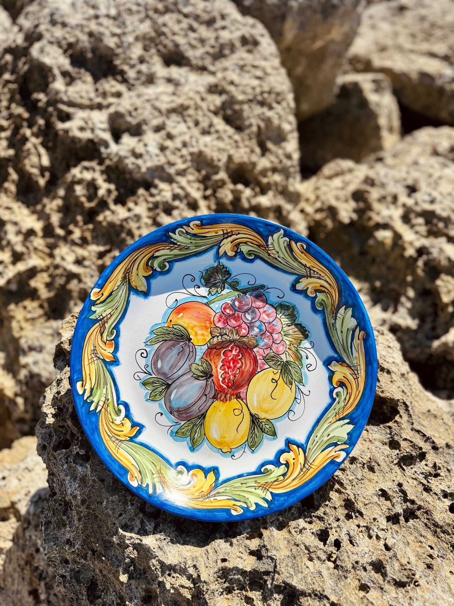 Handcrafted Sicilian ceramic fruit plate resting on a sandy Mediterranean beach, featuring colorful fruit motifs and traditional Caltagirone artisan decoration.