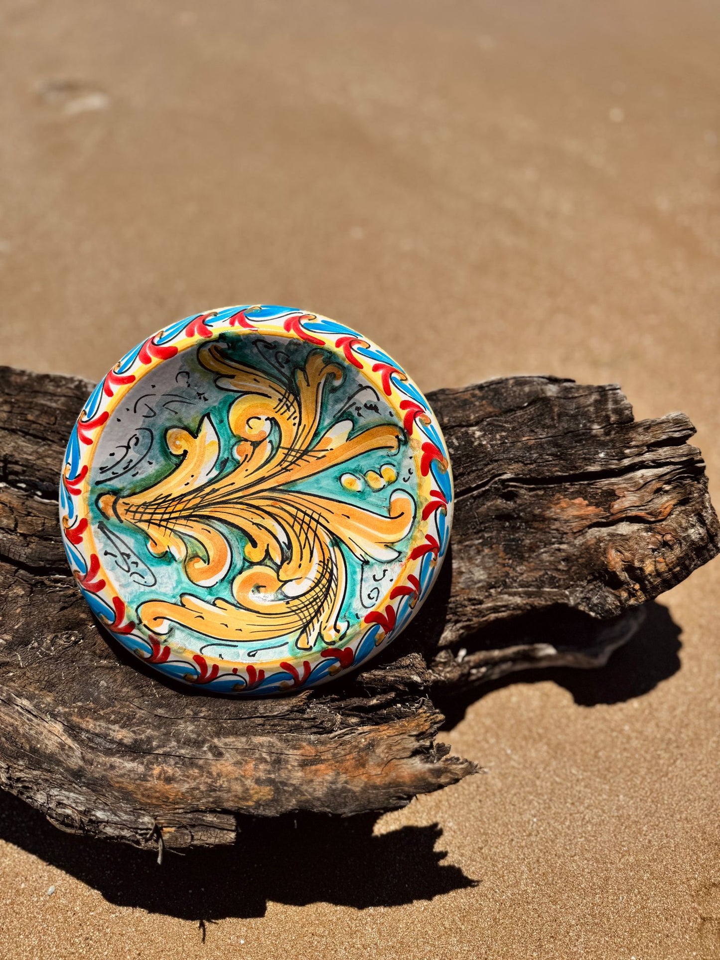 Hand-painted Sicilian majolica ceramic bowl resting on driftwood at the beach, inspired by Mediterranean craftsmanship and coastal living.