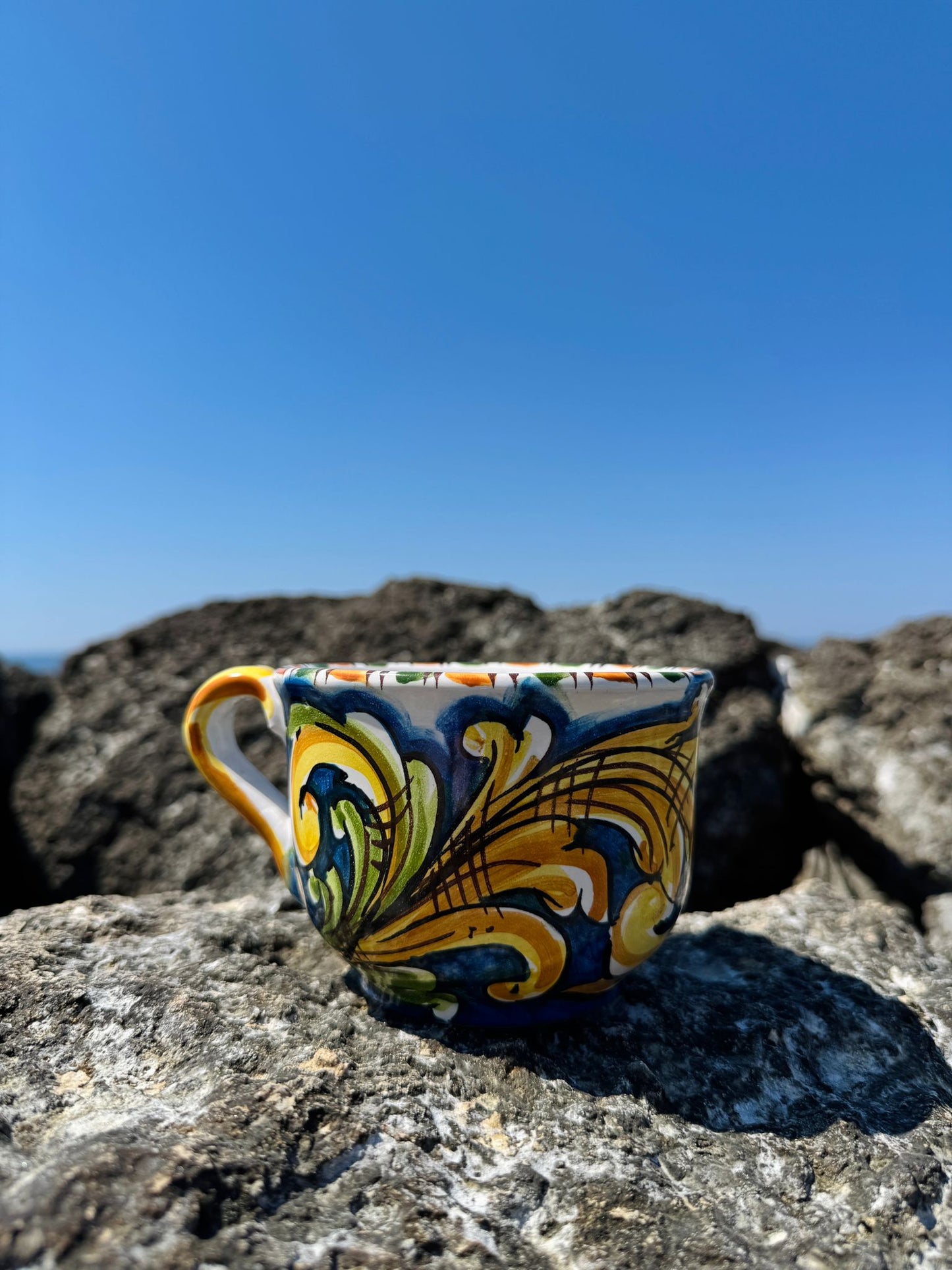Handcrafted Sicilian ceramic cappuccino cup with blue majolica decoration displayed on coastal rocks overlooking the Mediterranean Sea.