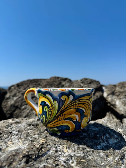 Handcrafted Sicilian ceramic cappuccino cup with blue majolica decoration displayed on coastal rocks overlooking the Mediterranean Sea.
