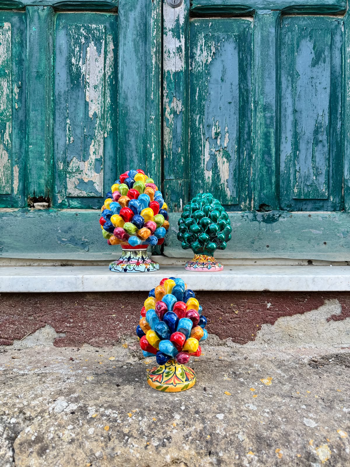 Three colorful decorative tree sculptures displayed outdoors against a weathered teal wooden door, with one larger multicolored tree in the foreground and two smaller green and multicolored trees behind it.