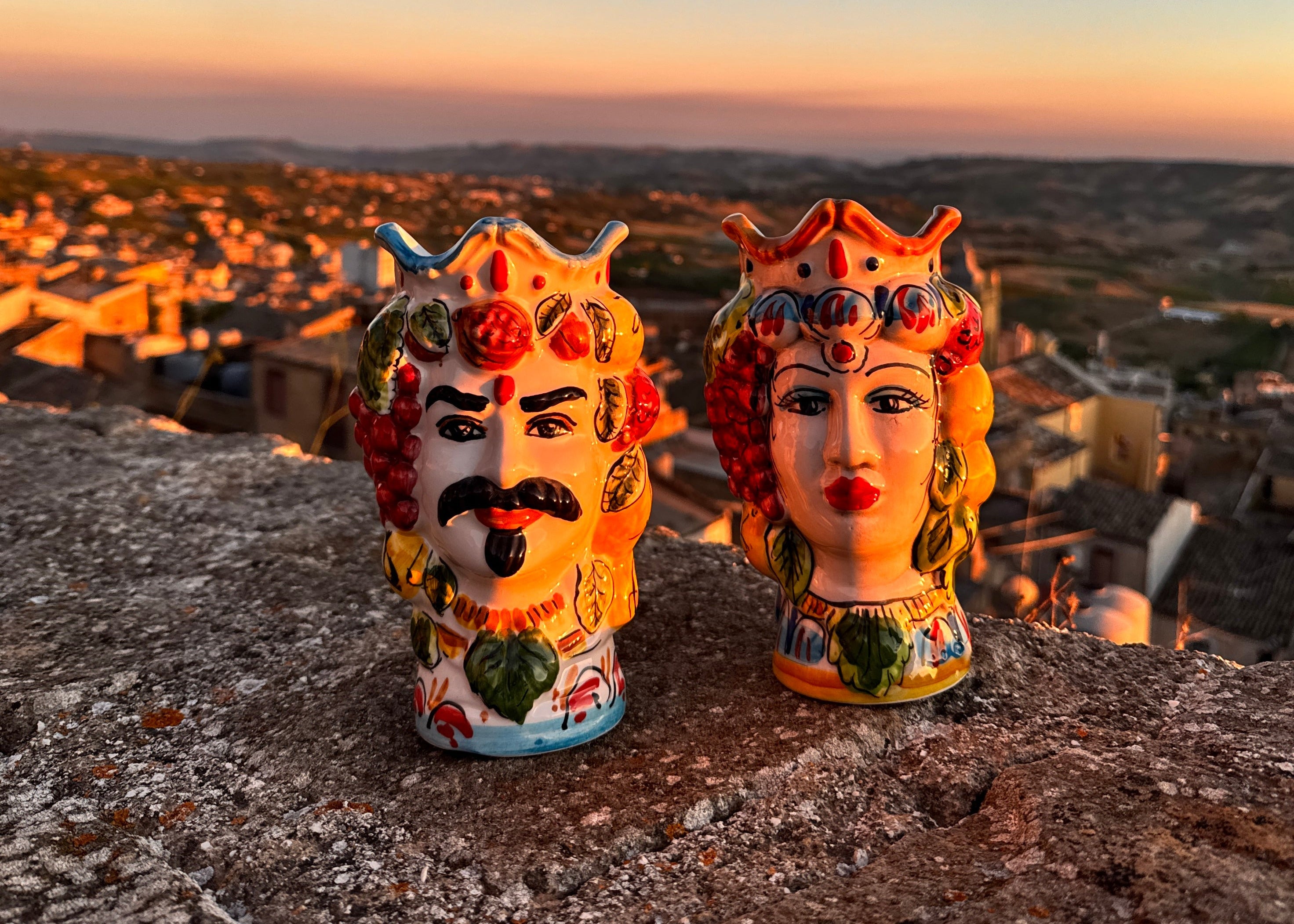 Two hand-painted ceramic salt and pepper shakers shaped like a king and queen, displayed on a stone ledge with a scenic sunset view of a town in the background.