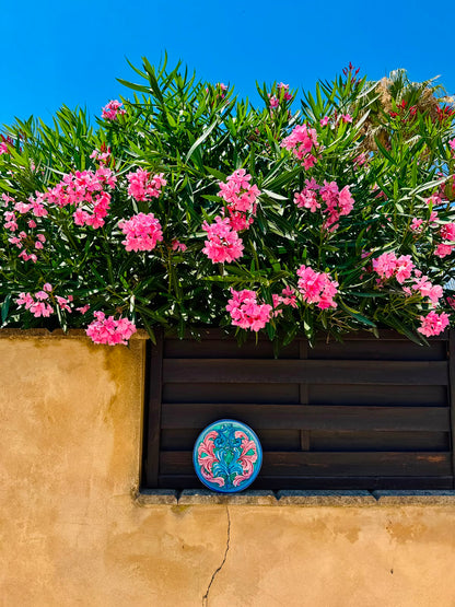 Hand-painted Sicilian ceramic plate displayed against a Mediterranean wall beneath pink oleander flowers, featuring turquoise, blue and pink majolica decoration.
