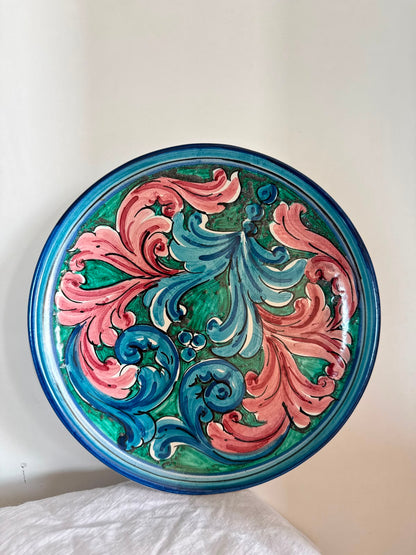 Hand-painted Sicilian ceramic decorative plate with pink, blue and green baroque motifs, displayed on natural white linen to highlight its artisan details.