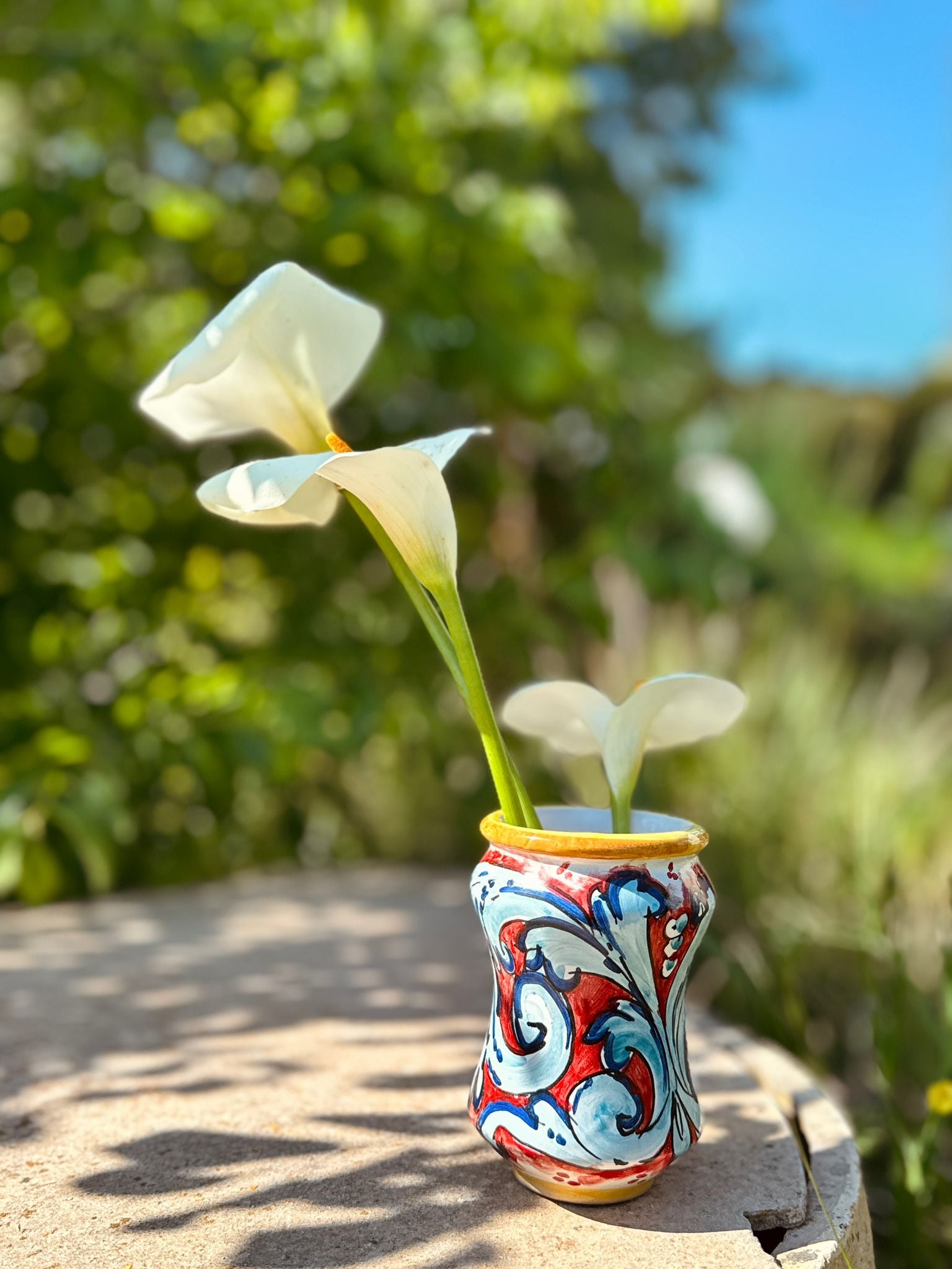 Handcrafted Sicilian ceramic flower vase with colorful majolica patterns and white calla lilies displayed outdoors in warm Mediterranean sunlight.