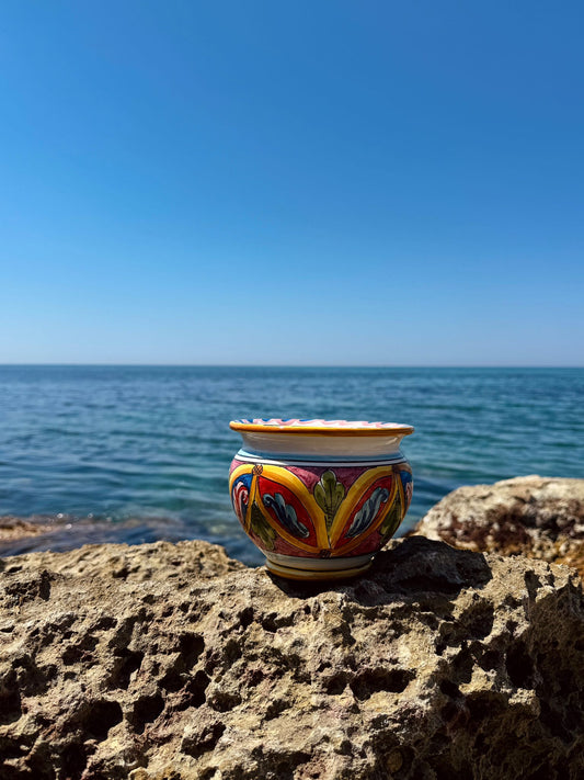Handcrafted Sicilian ceramic cachepot displayed on coastal rocks overlooking the Mediterranean Sea, featuring traditional Caltagirone majolica patterns.