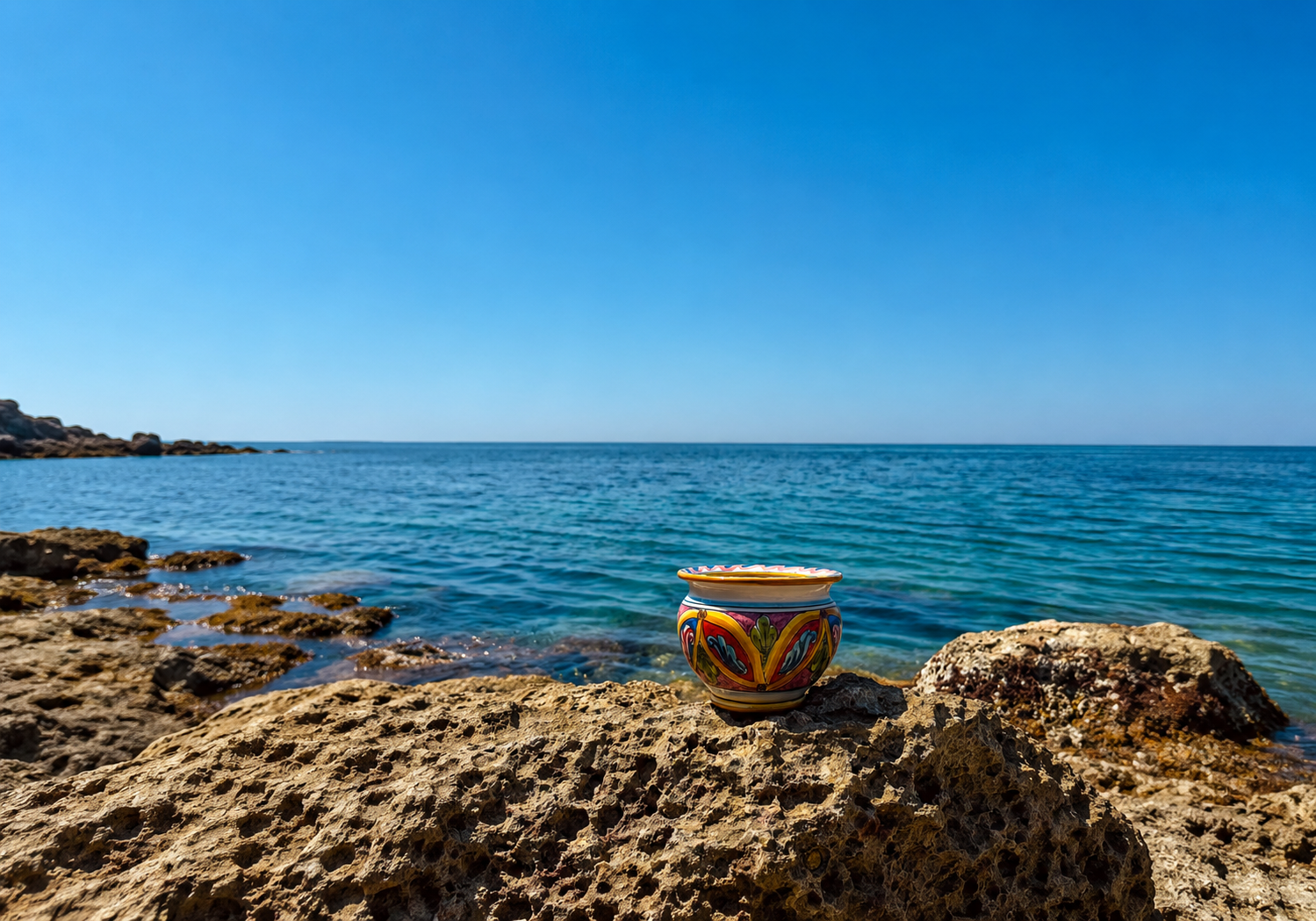 Handcrafted Sicilian ceramic cachepot displayed on coastal rocks overlooking the Mediterranean Sea, featuring colorful traditional majolica decoration.