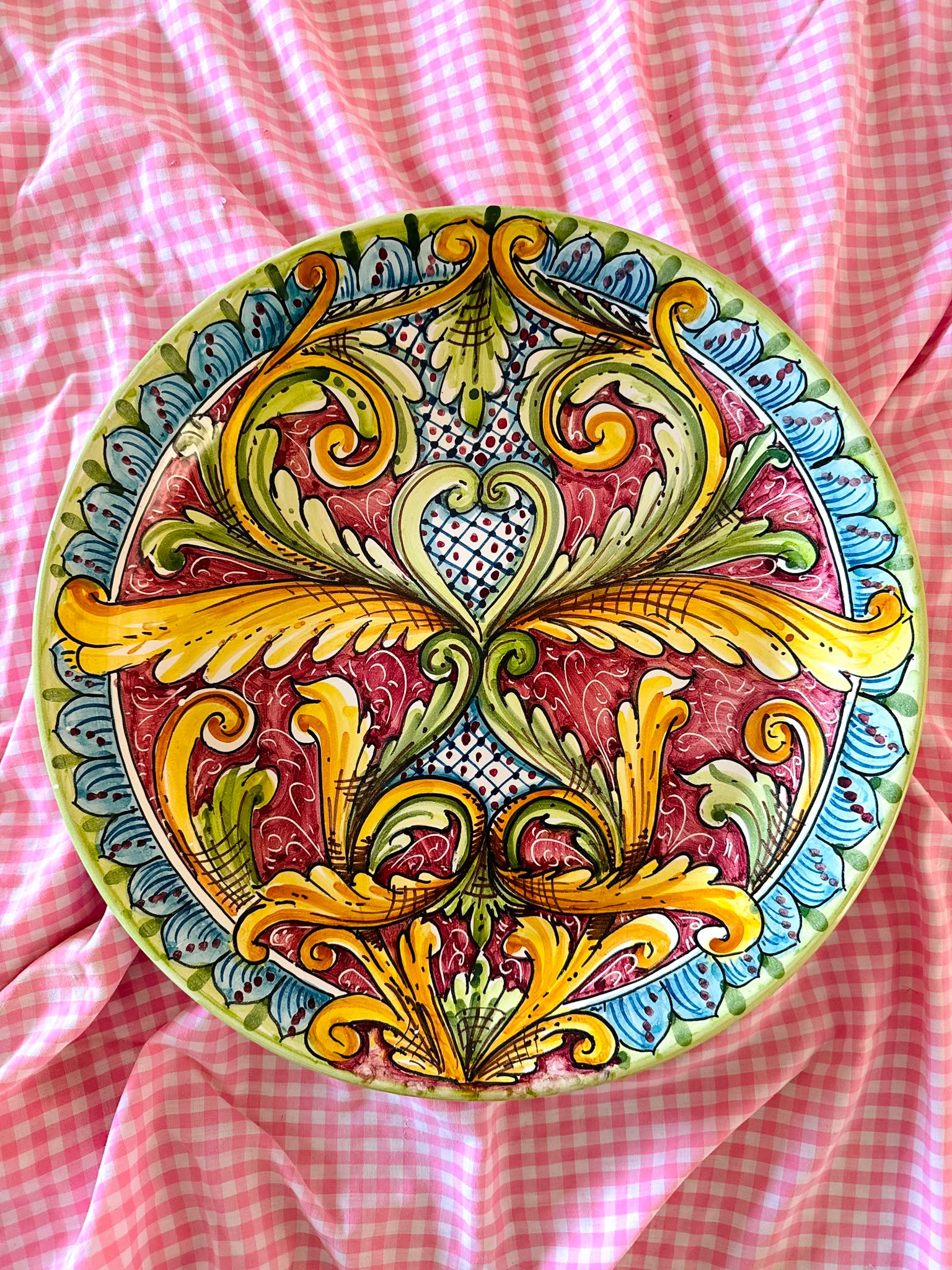 Hand-painted Sicilian ceramic decorative plate with burgundy, yellow, green and blue baroque majolica decoration, displayed on pink gingham fabric.