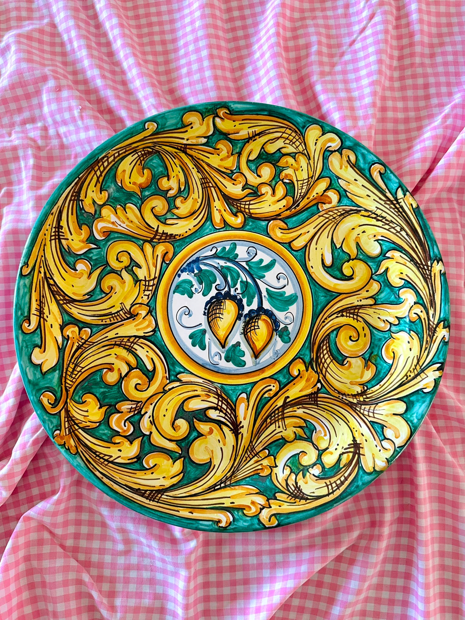Hand-painted Sicilian ceramic decorative plate with green and gold baroque majolica decoration and central ornamental motif, displayed on pink gingham fabric.