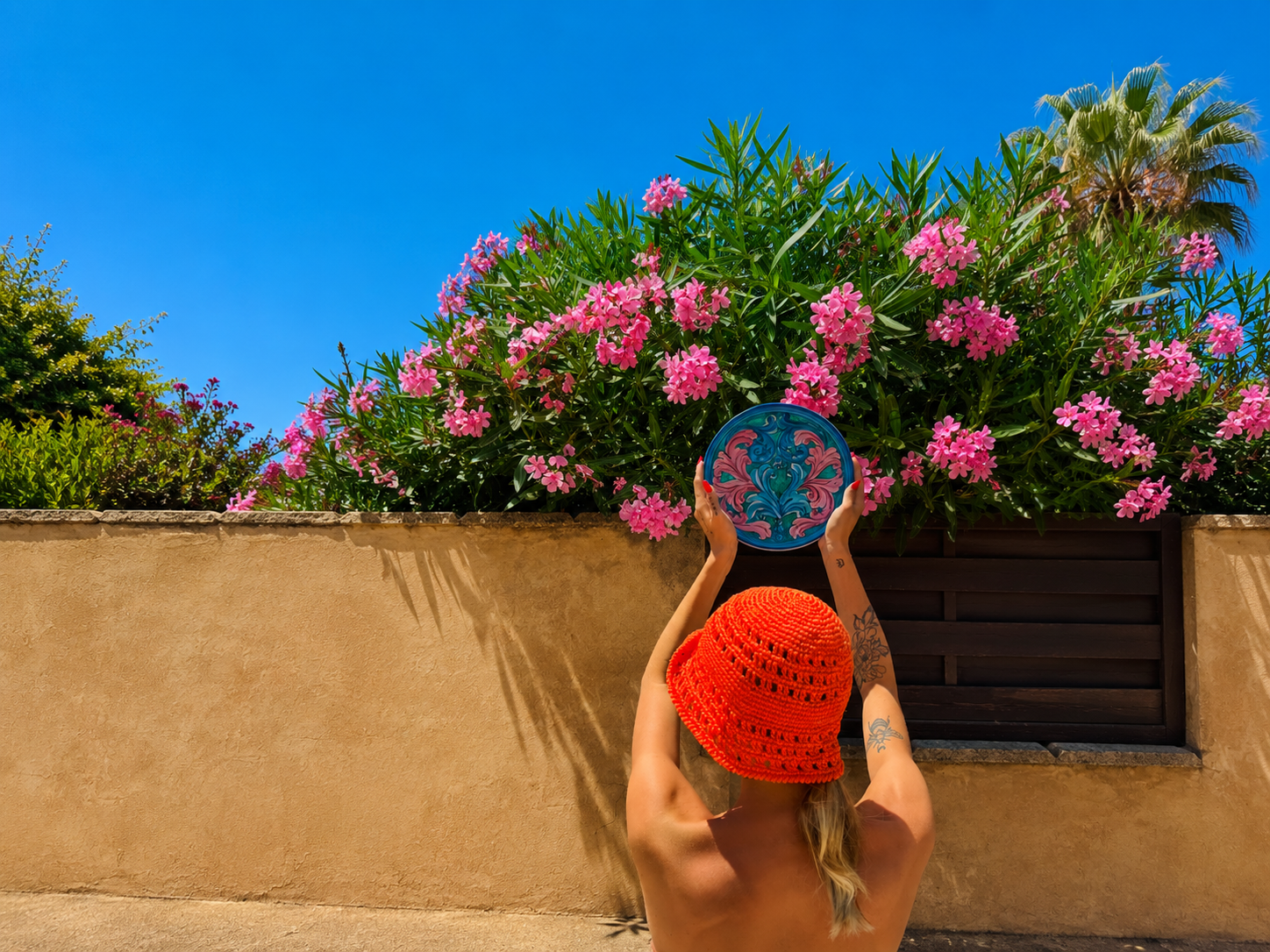 Handcrafted Sicilian ceramic decorative plate with pink and blue baroque majolica decoration, held in a sunny Mediterranean garden with pink flowers.