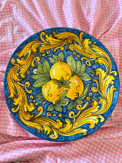 Hand-painted Sicilian ceramic decorative plate featuring yellow lemons, green leaves and blue baroque majolica motifs, displayed on pink gingham fabric.