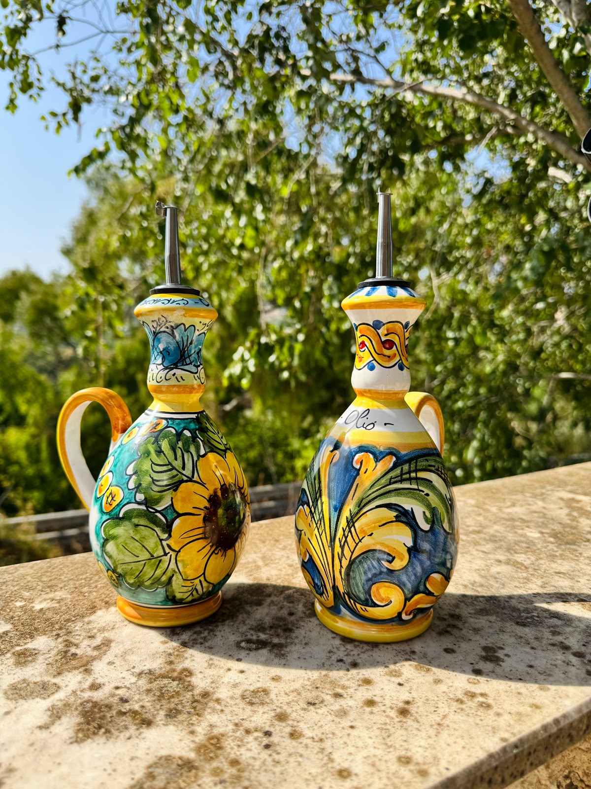 Pair of hand painted Sicilian ceramic olive oil bottles displayed outdoors in a Mediterranean garden, featuring colorful Caltagirone inspired majolica decoration.