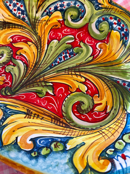 Hand-painted Sicilian ceramic oval platter with colorful baroque majolica decoration, displayed on pink gingham fabric to highlight its Mediterranean character.