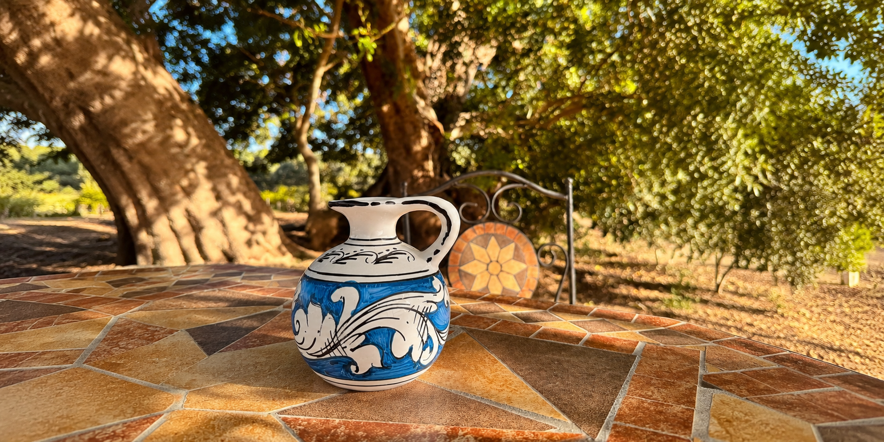 Handcrafted Sicilian ceramic pitcher with blue and white majolica patterns displayed on a rustic Mediterranean mosaic table surrounded by olive trees and warm Sicilian sunlight.