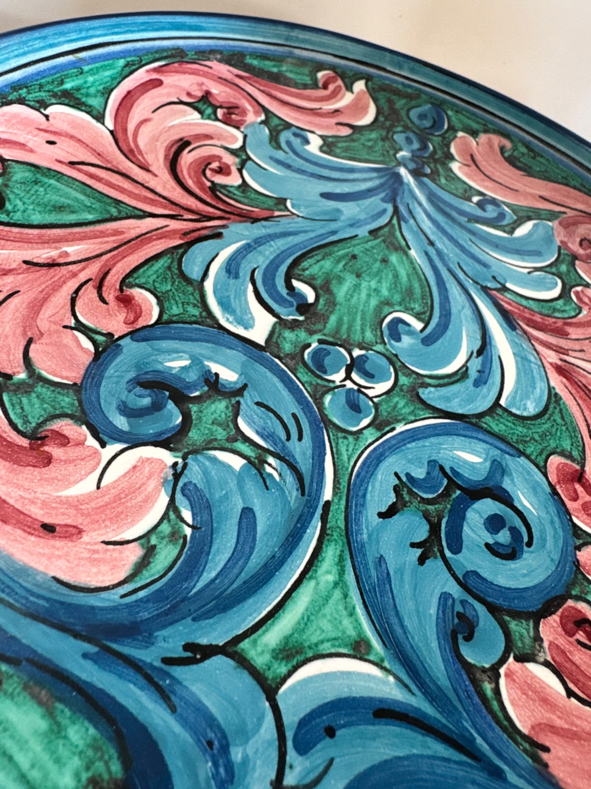 Close-up of a handcrafted Sicilian ceramic plate showing glossy pink, blue and turquoise baroque majolica brushwork and artisan hand-painted details.