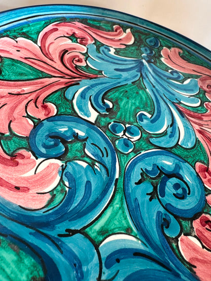 Close-up of a handcrafted Sicilian ceramic plate showing glossy pink, blue and turquoise baroque majolica brushwork and artisan hand-painted details.