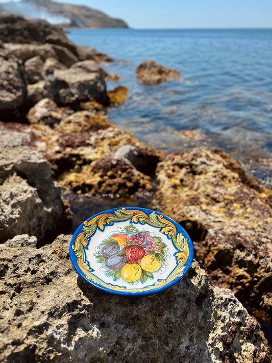 Handcrafted Sicilian ceramic fruit plate displayed on natural Mediterranean rocks, featuring lemons, figs, grapes and pomegranate with traditional Caltagirone decorations.