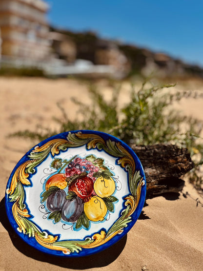 Handcrafted Sicilian ceramic fruit plate displayed on seaside rocks overlooking the Mediterranean Sea, inspired by traditional Caltagirone majolica craftsmanship.