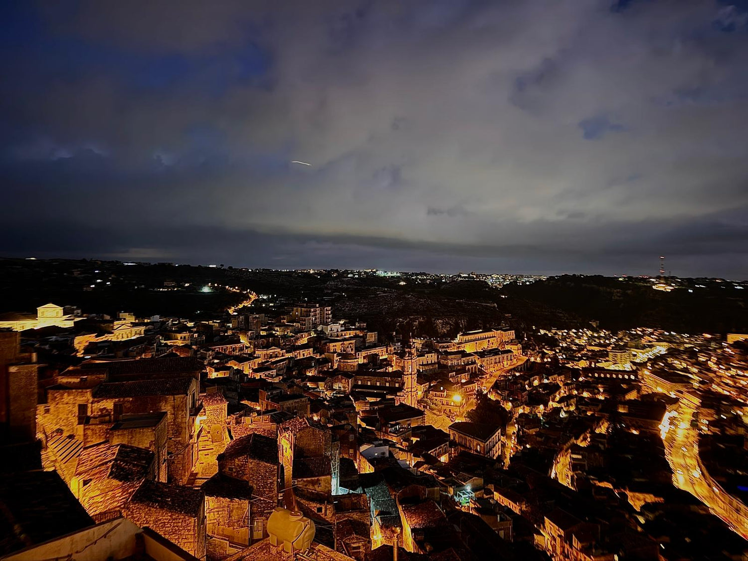 Panoramic nighttime view of a historic Sicilian town illuminated by warm golden lights under a dramatic Mediterranean sky.