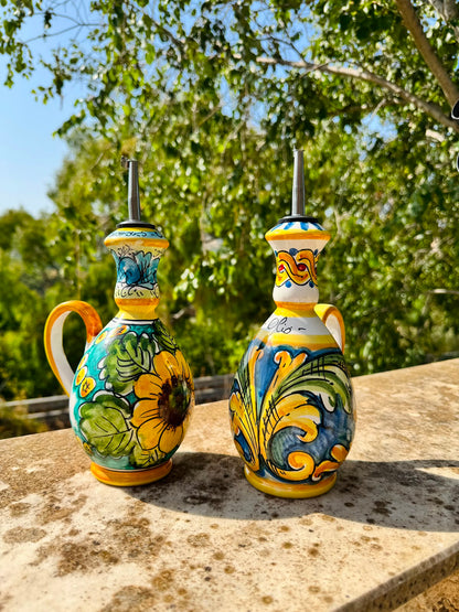Pair of hand painted Sicilian ceramic olive oil bottles displayed outdoors in a Mediterranean garden, featuring sunflower and baroque majolica decoration.
