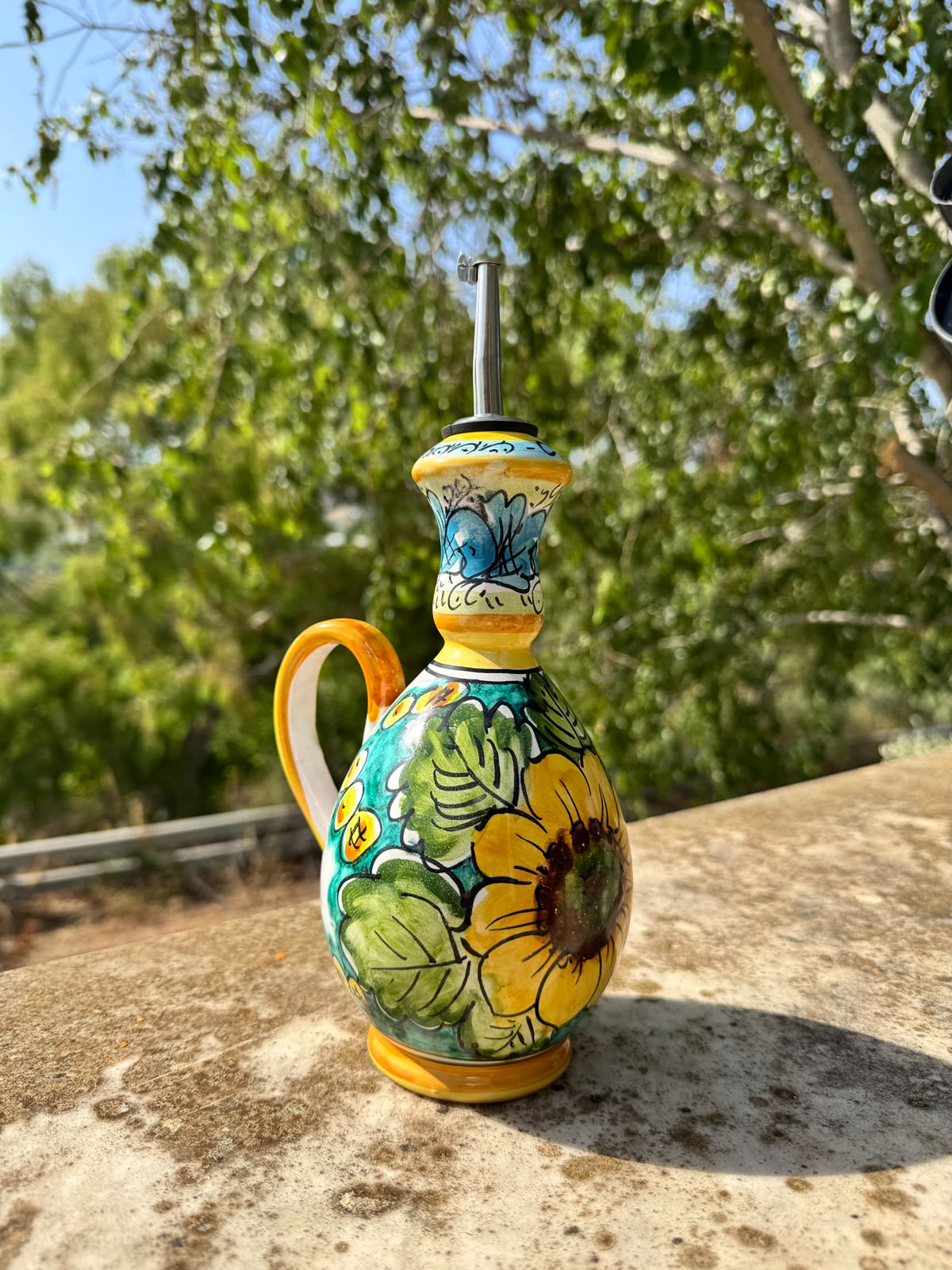 Handcrafted Sicilian ceramic olive oil bottle with sunflower motif, photographed outdoors in a sunny Mediterranean garden.
