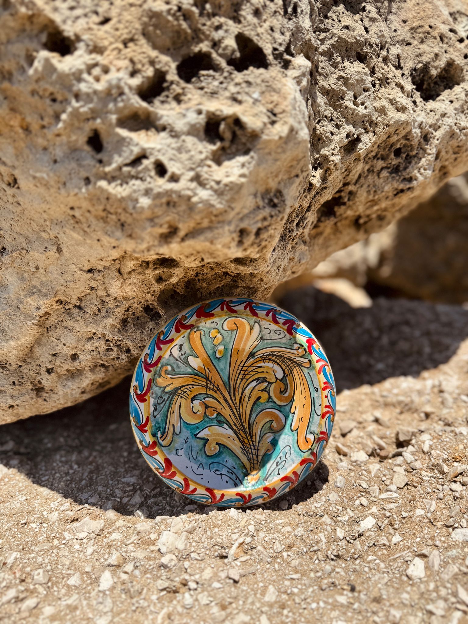 Handcrafted Sicilian ceramic bowl displayed among Mediterranean coastal rocks, showcasing traditional Caltagirone baroque decoration.