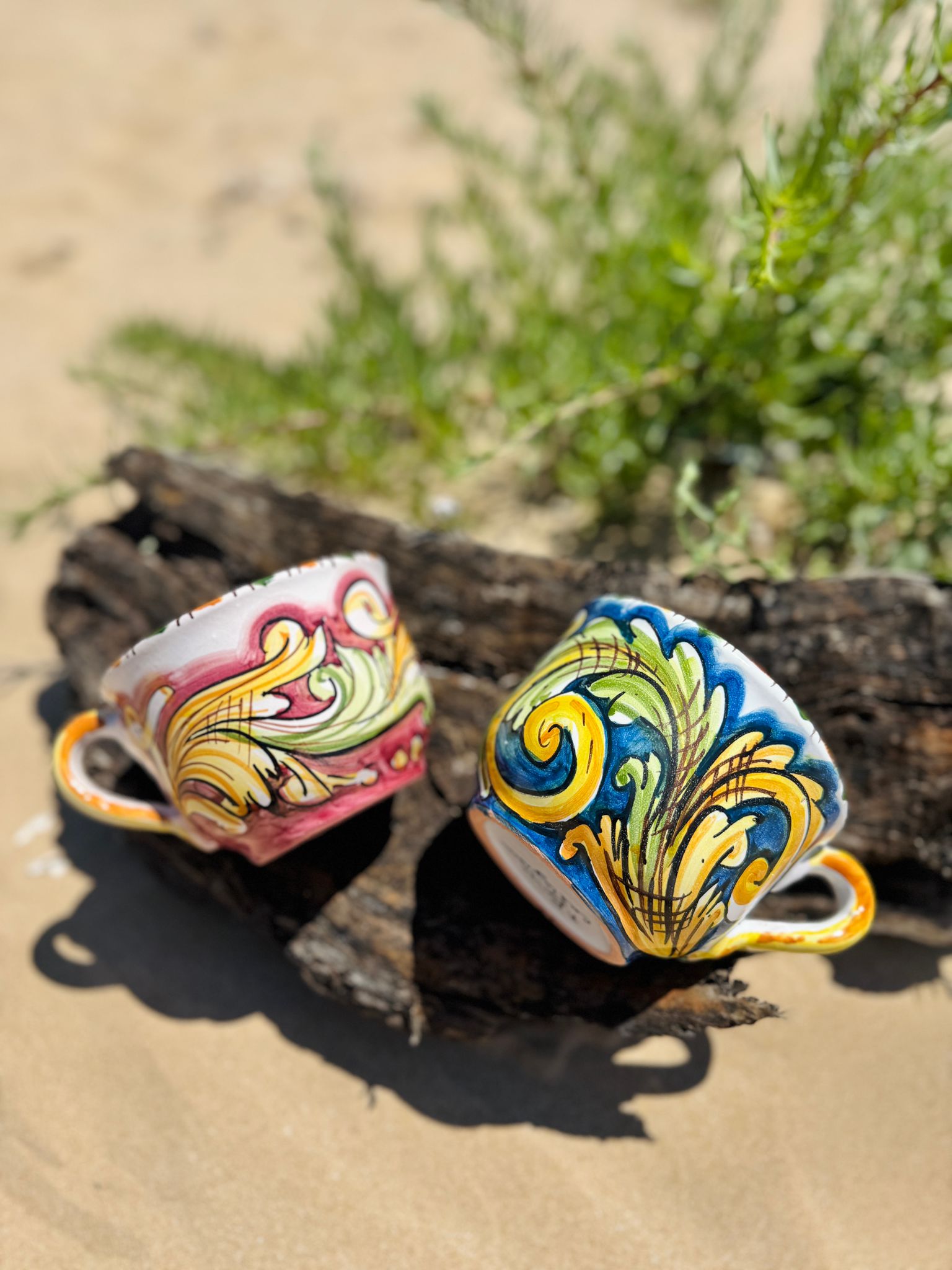 Pair of handcrafted Sicilian ceramic cappuccino cups resting on driftwood along a Mediterranean beach, showcasing traditional majolica patterns.