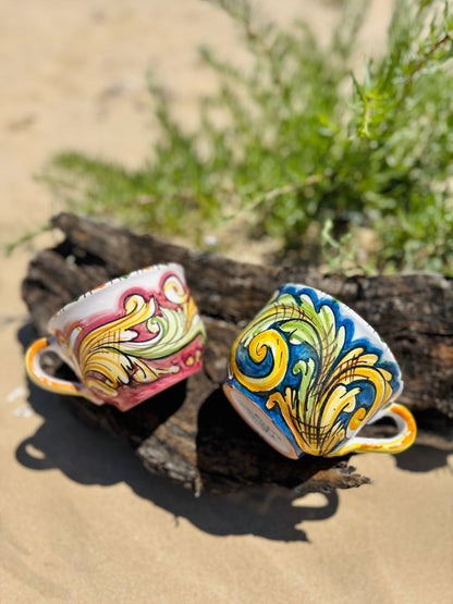 Pair of handcrafted Sicilian ceramic cappuccino cups resting on driftwood along a Mediterranean beach, showcasing traditional majolica patterns.