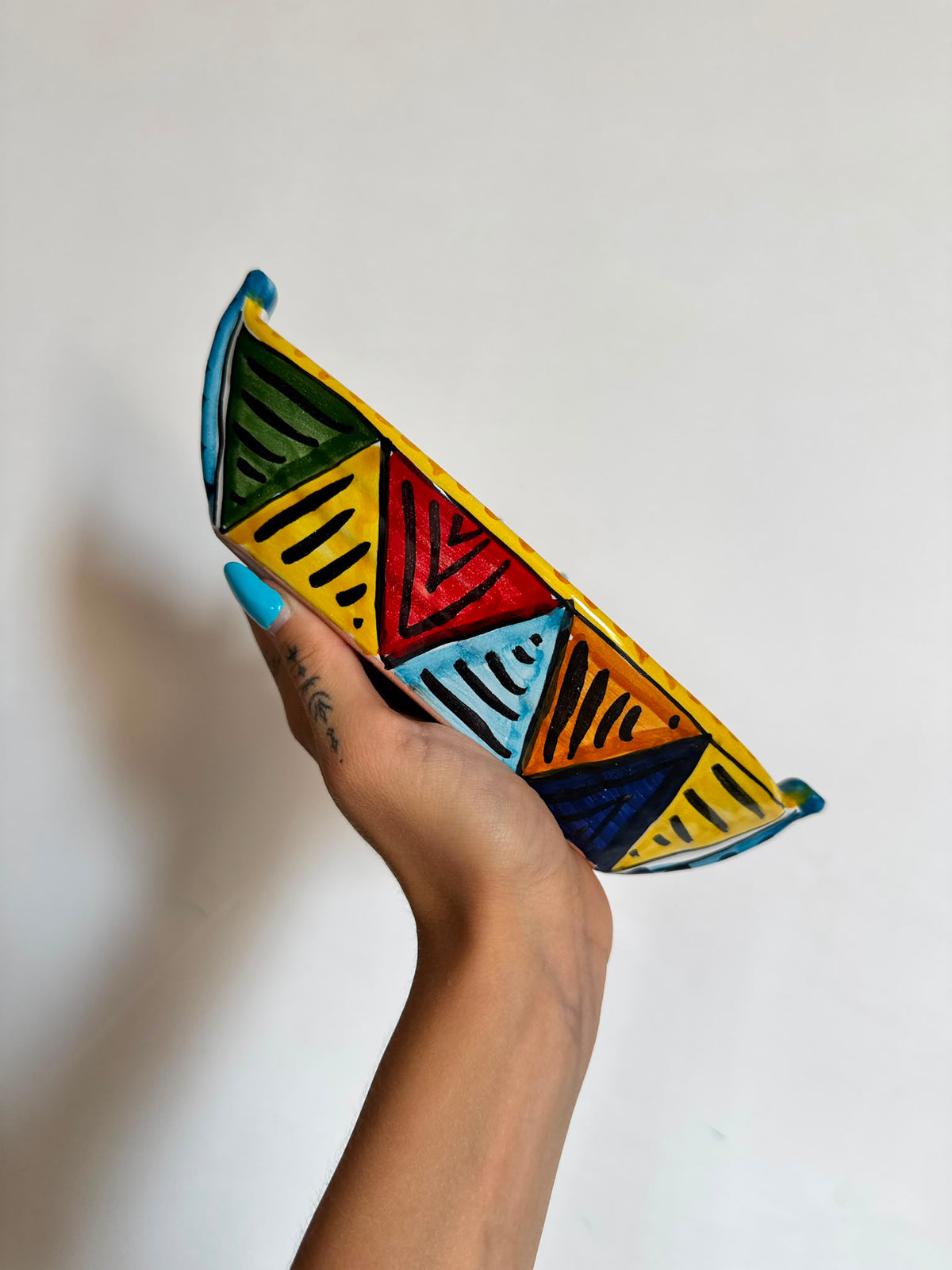 A hand with blue nail polish holds a small hand-painted ceramic bowl featuring a colorful geometric pattern of triangles in yellow, green, red, light blue, and dark blue against a plain white background.